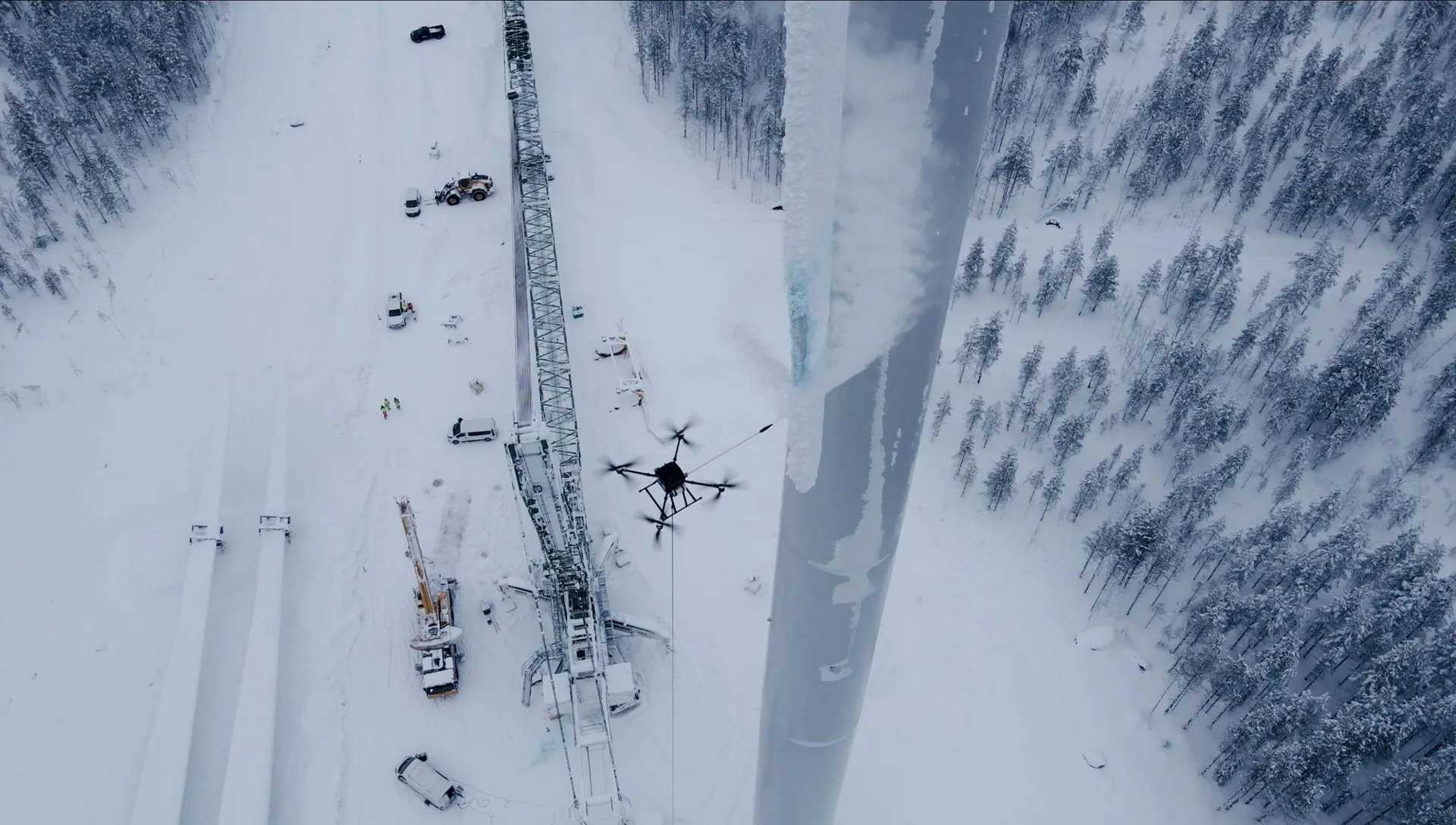 Drone ved en isdækket vindmøllevinge med kran og køretøjer på den snedækkede arbejdsplads